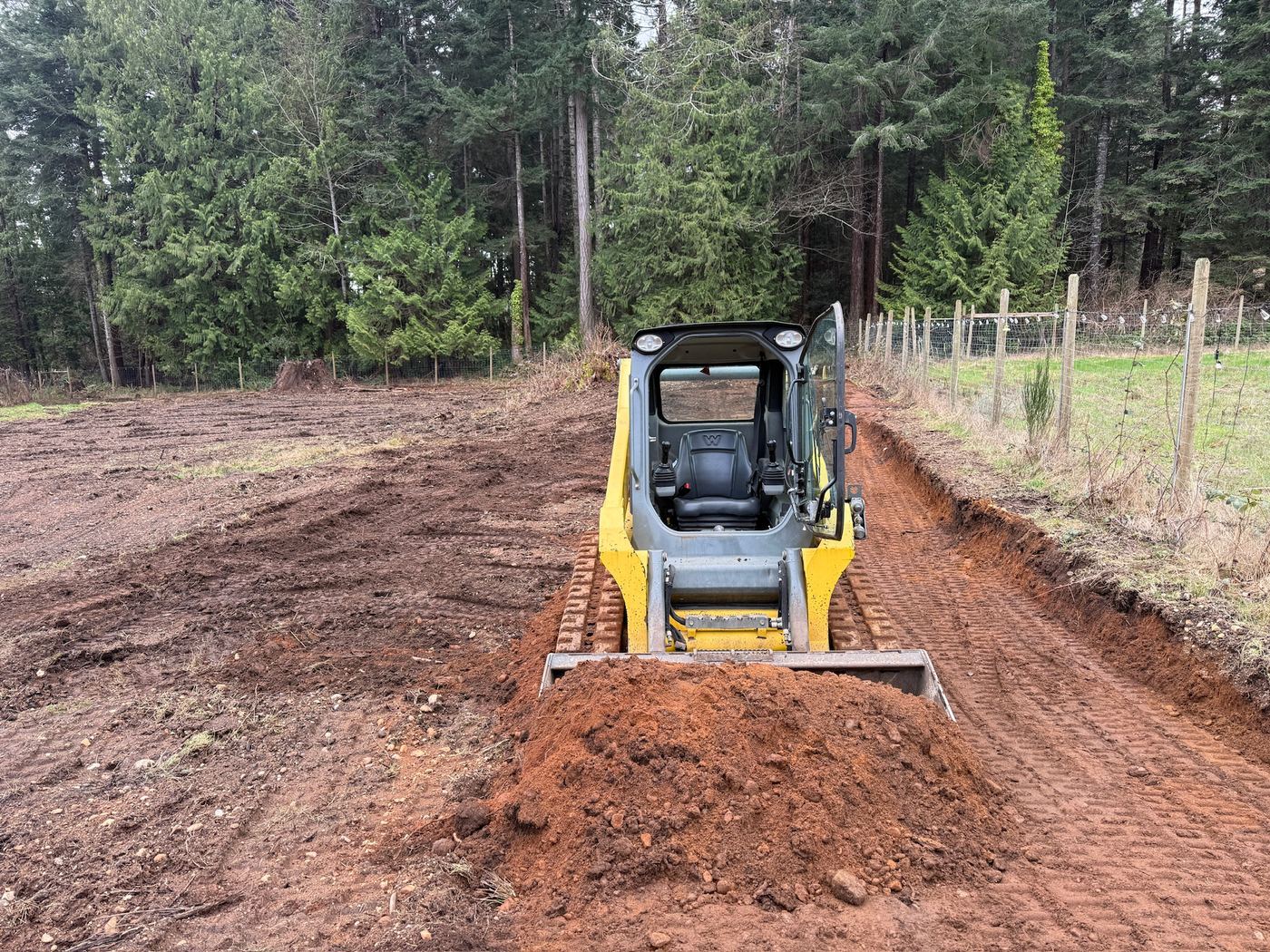 A track loader with a bucket of red soil on the new road